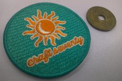 エメグリーンのクロス生地に、オレンジ、薄黄の糸で暑そうな太陽のイラストと企業名を刺繡した、オリジナル刺繡ワッペンの制作実績。