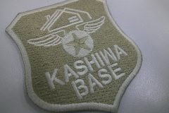 ベージュ系のクロス生地にクリーム系の糸で企業ロゴを刺繡し、盾形のオリジナル刺繡ワッペンの制作実績。
