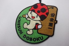 緑色の生地に、テントウムシが安全第一の看板を掲げて歩くイラストのオリジナル刺繡ワッペンの制作実績。