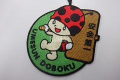 緑色の生地に、テントウムシが安全第一の看板を掲げて歩くイラストのオリジナル刺繡ワッペンの制作実績。
