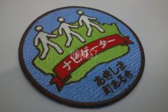 水色の生地に緑色で昭島を描き、その上を白糸で三人が歩いているイラストと団体名と刺繡した、オリジナル刺繡ワッペンの制作事例。