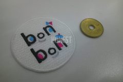 文字を顔に見立ててピンクや水色で目や装飾を刺繡した、白い生地の円形のオリジナル刺繡ワッペンの制作事例。