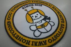 白い生地にオレンジ色の糸で蜂がモチーフのキャラクターと団体名などを刺繡した、オリジナル刺繡ワッペンの制作事例。