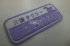 白のツイル生地に薄めのバイオレットカラーで鴨のシルエットと併せ、介護タクシーの社名を刺繍した制作事例。こちらは横長タイプでの制作。