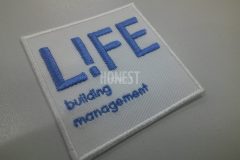 白のツイル生地をベースに、サックスブルーで建設会社のロゴを刺繍した、シンプルなオリジナルワッペンの制作事例。