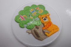 白い生地にオレンジのクマが木に抱き着いたイラストの葉っぱにカラフルな糸で団体名を刺繡したオリジナル刺繡ワッペンの制作実績。