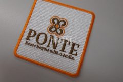 白い生地にオレンジとブラウンの糸で店名とロゴを刺繡した、オリジナル刺繡ワッペンの制作実績。