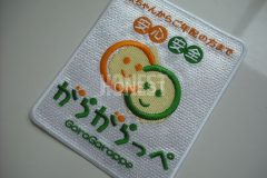 白色のクロス生地に緑色とオレンジ色の糸で刺繡したオリジナル刺繡ワッペンの制作事例。