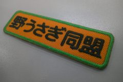 濃いめのオレンジのツイル生地に文字は黒糸。外周フチはケリーグリーンの糸で仕上げた、文字ワッペンの加工事例。