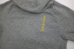 グレーのパーカーの背面上部の右よりに金糸で縦方向に黄色の刺繍文字が加工された制作事例。
