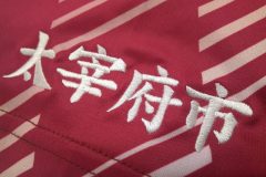 エンジ色の競技チームのユニフォームシャツの右袖に白糸で文字刺繍をした制作事例のアップ。