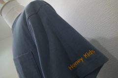 お持ち込みのTシャツ左袖口に濃いめのオレンジ糸で保育園名を英字で加工した制作実績。書体はRound Blockを使用。