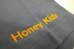 お持ち込みのTシャツ左袖口に濃いめのオレンジ糸で保育園名を英字で加工した制作実績の平置きアップ。