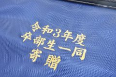 部活動の卒部記念の刺繍の製作実績。青のナイロンバッグに金糸、楷書体で文字を三段構成で刺繍した。