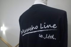 黒い長袖シャツの背面に白い筆記体の企業ロゴ。下にブラシストローク風のラインが入ったシンプルでスタイリッシュに仕上げたフルカラー転写プリントの制作事例。