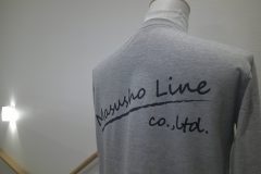 グレーの長袖シャツの背面に黒い筆記体の企業ロゴ。下にブラシストローク風のラインが入ったシンプルでスタイリッシュに仕上げたフルカラー転写プリントの制作事例。
