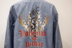 淡いブルーデニムシャツの背面に、金と黒のトライバル模様と赤いゴシック文字『Voluntad de luchar』が加工されたフルカラー転写プリントの制作事例。