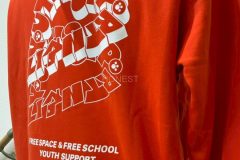 オレンジ色のプルジップパーカーの背中全面に白いシートで団体様のイメージをデザインされたイラストとロゴマークをプリントしたラバープリント部分のアップ。