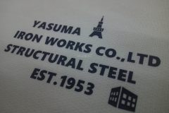 アイボリーの裾ドローコード付きTシャツの左胸にネイビーのシートで、社名や取扱い業務、創業年などをデザインしたマークをプリントしたラバープリント部分のアップ。