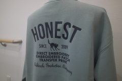 ミントグレーのトレーナーの背中全面に黒いシートで大きく「Honest」のロゴを配置したラバープリントのアップ。