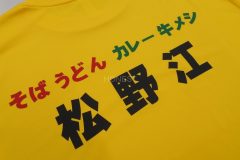 黄色いTシャツの背中中央に黒いシートで飲食店名、赤、緑のシートでメニューをデザインしたラバープリントの制作見本。