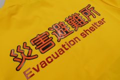 黄色いポロシャツの背中中央に災害避難所とプリントしたラバープリント部分のアップ。下に黒いシート、上にオレンジ色のシートを重ねてプリント圧着することによって囲み文字を表現している。