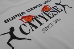 白いTシャツの胸中央にダンスユニット名をプリントしたラバープリント部分のアップ。赤、オレンジ色のシートで瞳のイラスト。黒いシートで文字と人物のシルエットを。シートを重ねプリントすることで表現している。
