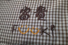 チェック柄のチュニックの左胸にブラウンとオレンジの糸で企業のロゴを刺繍加工した制作実績のアップ。