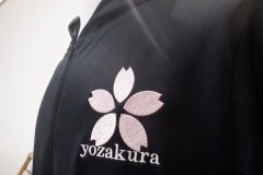 パーカー左胸に淡いピンクの糸で桜のマーク。その下に英字で会社名を刺繍した制作事例。