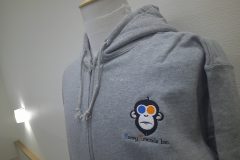 グレーのフルジップパーカー左胸に白・橙・青・黒で企業ロゴをオリジナル刺繍として加工した制作実績。