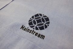 オックスフォードシャツの左胸に企業ロゴを刺繍。ネイビーのロゴマークはタタミ縫い。黒文字はサテン縫いで仕上がり印象に緩急をつけた制作実績。