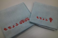 薄い水色のハンドタオル2枚にそれぞれ企業ロゴとトラックのモチーフと企業ロゴと気球のモチーフを赤糸で刺繍した制作実績。