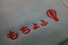 薄い水色のハンドタオルに企業ロゴと気球のモチーフを赤糸で刺繍した制作実績。