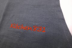 炭黒のエプロンに赤糸でオリジナルロゴ文字が刺繡された制作事例のアップ。