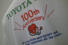 白いポロシャツの背面に緑・赤・青の糸で企業の100周年を祝うためのオリジナルロゴ刺繍の制作事例の右サイドからのアップ。