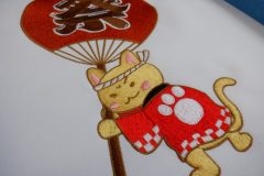白字に赤・白・黒・茶・グレー・肌色の糸でお祭りを楽しむ猫が表現されたオリジナル刺繍の制作事例。