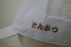 帽子左側面に勘亭流書体で「とんかつ」とシンプルに文字刺繍をした事例。糸の色はブラウン。
