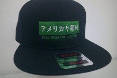 黒の帽子の前面にグリーンのボックスロゴ。その下に地域名を刺繍した制作実績。