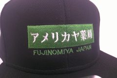 黒の帽子の前面にグリーンのボックスロゴ。その下に地域名を刺繍した制作実績のアップ。