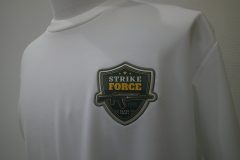 白いTシャツの左胸にミリタリー風のイラストがプリントされエンブレム型生地をベージュの糸で縁取り刺繡した制作見本。