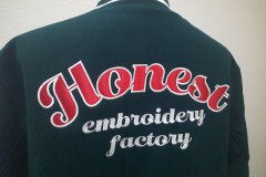 メルトンジャケットの背面にアーチ状の縁取り刺繍を加工し、その下に英字（筆記体）を直刺繍した作品のアップ。