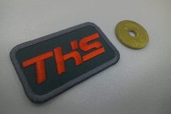 特徴的な書体で会社名を刺繍した文字ワッペン。文字色はオレンジ。生地はチャコールカラー。