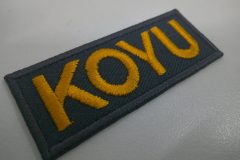 チャコールのツイル生地をベースにオレンジの糸で企業名を刺繍した、ミニサイズの文字ワッペンの制作実績。