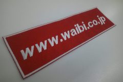 赤の生地を土台に、ブロック体を斜体に調整した企業のURLを白糸で刺繍した文字ワッペンの制作実績。