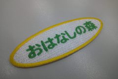 白のエンブクロス生地に緑の糸で福祉施設名を刺繍。外周フチは黄色の糸。可愛らしい丸ゴシック体で製作。