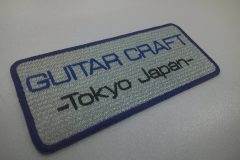 シルバーグレーのエンブクロス生地に青糸と黒糸でギタークラフト工房の名前を刺繍した、文字ワッペンの制作事例。