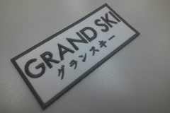 白のツイル生地にスポンサー名をグレー糸で刺繍した、文字ワッペンの制作事例。二段構成。