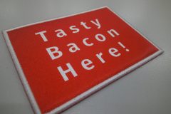 赤い長方形に白い文字で『Tasty Bacon Here!』と書かれたオリジナルプリントワッペンの制作見本。」