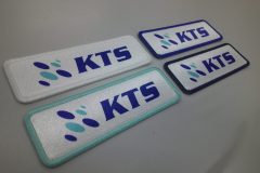 白または銀地に青い“KTS”ロゴが入った長方形のオリジナルプリントワッペン4枚の制作実績。左側に青とターコイズの楕円形ロゴがあり、縁取りの色が白・ターコイズ・濃紺で異なる。