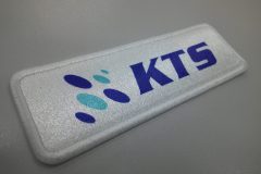 白地に青い“KTS”ロゴと楕円形マークが加工された長方形オリジナルプリントワッペンの制作事例。